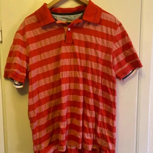 Röd och rosa randig pikétröja från Hugo Boss - Säljer en snygg röd och rosa randig pikétröja från Hugo Boss. Tröjan har en klassisk krage och korta ärmar, perfekt för en avslappnad stil. Regular fit för en bekväm passform.