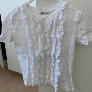 Jättesöt vit blus-T-shirt från Zara ☀️ - Säljer denna söta vita blus / T-shirt  från Zara med volangdetaljer framtill. 🤍 Tröjan är i samma material som en vanlig T-shirt men ser ut som en blus framtill. Har används fåtal gånger, köptes second hand 🌸 Skriv vid mer frågor / bilder 💛Märke: Zara, Storlek:Xs-S