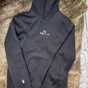 Marinblå Hoodie från Ralph Lauren - Snygg marinblå hoodie från Ralph Lauren med broderad logga på bröstet och den klassiska polospelaren på sidan. Tröjan har en stor ficka framtill och justerbar huva med snören. Perfekt för en stilren och bekväm look. Nypris 2100kr, Vårat pris 800kr! Priset kan diskuteras.