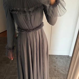 Grå långklänning med volangdetaljer - Elegant grå långklänning i ett lätt och luftigt material med fina volangdetaljer vid ärmslut och midja. Klänningen har långa ärmar och en mjuk, flytande passform. Är 166cm 