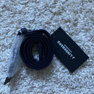 Blått flätat bälte från J.Lindeberg - Säljer ett snyggt blått flätat bälte från J.Lindeberg i nytt skick. Bältet är 85 cm långt och har en stilren design med läderdetaljer. Perfekt för att ge din outfit en sportig och elegant touch. Passar både till vardags och fest! 700 kr nypris.