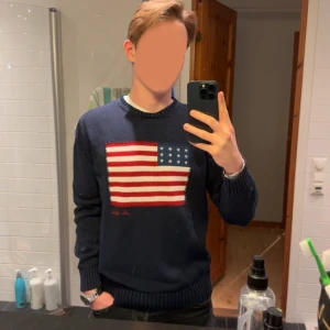 Mörkblå tröja med amerikansk flagga - Säljer en snygg mörkblå tröja från John Ashfield med en stor amerikansk flagga på framsidan. Tröjan är i bra skick och har långa ärmar med ribbade muddar. Perfekt för höst och vinter!