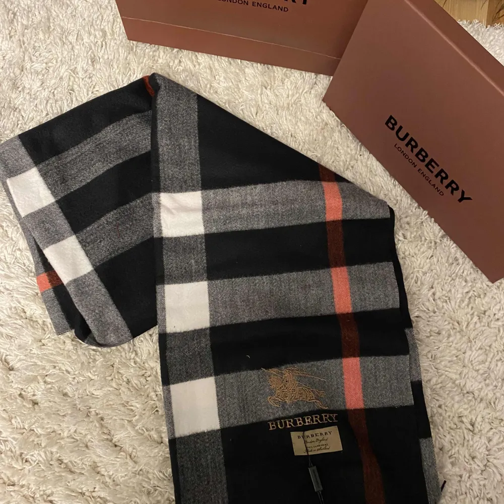 Säljer en superfin rutig halsduk från Burberry i klassiska färger som svart, grå och vit med en touch av röd. Den är i nytt skick och kommer med originalförpackning. Perfekt för att hålla sig varm och stilren under höst och vinter. 🧣✨. Övrigt.