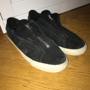 Svarta sneakers från Axel Arigato - Säljer ett par svarta sneakers från Axel Arigato i mocka med dragkedja framtill. De har en vit sula som ger en snygg kontrast. Perfekta för både vardag och fest! Skorna är i bra skick med lite slitage på sulan.