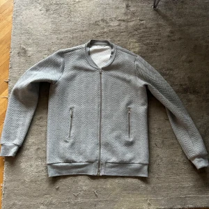 Grå tröja från J.Lindeberg - Säljer en snygg grå bomberjacka från J.Lindeberg i mycket bra skick. Jackan har ett coolt sicksackmönster och dragkedja framtill. Perfekt för höst och vår! Den har långa ärmar och ribbade muddar vid ärmslut och nederkant. Passar både till vardags och lite finare tillfällen.
