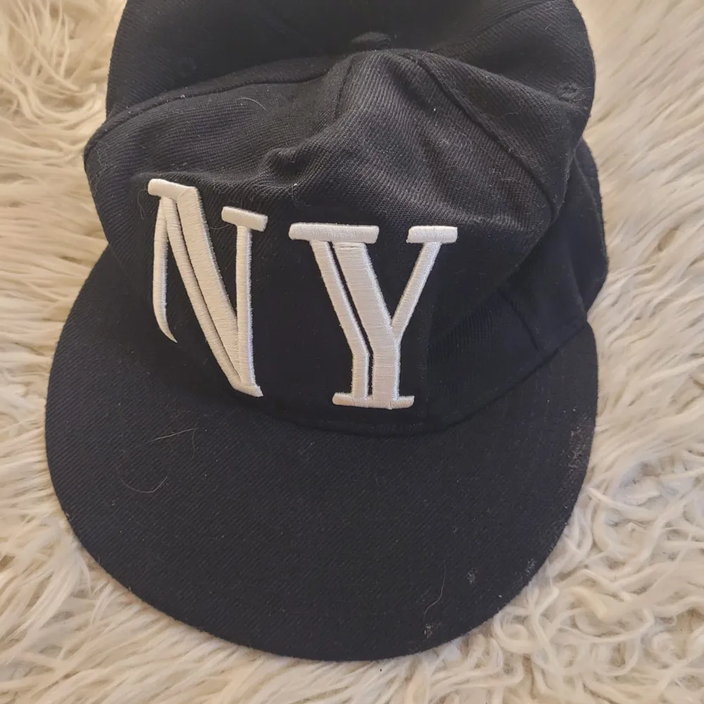 Snygg svart keps med stort vitt 'NY' broderat framtill och 'NEW YORK' på undersidan av skärmen. Tillverkad i 80% bomull och 20% polyester, vilket gör den både bekväm och hållbar. Perfekt för en casual look eller för att visa din kärlek till New York!. Asusteet.