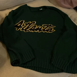 Grön stickad tröja från H&M Divided - Säljer en snygg grön stickad tröja från H&M Divided med texten 'Atlanta' i gult och svart på framsidan. Tröjan har långa ärmar och ribbade kanter. Perfekt för en avslappnad stil i höst och vinter.