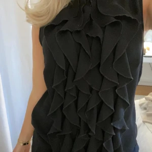 Svart ärmlös blus med volanger - Snygg svart ärmlös blus med volanger framtill. Perfekt för festliga tillfällen! Blusen  har en elegant och chic stil som passar både till vardags och fest. Materialet känns bekvämt och lätt att bära.