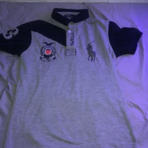 Grå och marinblå pikétröja från Polo Ralph Lauren - Säljer en snygg grå och marinblå pikétröja från Polo Ralph Lauren. Den har korta ärmar och en klassisk krage med knappar. På framsidan finns broderade detaljer och en stor logga. Perfekt för en avslappnad men stilren look!