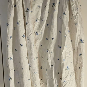 Vit blommig klänning från H&M - Säljer en superfin vit klänning från H&M med blå blommor. Kjolen har en elastisk midja för extra komfort och är perfekt för vår och sommar. Materialet känns lätt och luftigt, vilket gör den idealisk för varmare dagar. Passar både till vardags och festligare tillfällen!