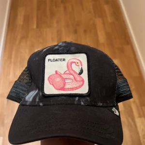 Svart keps med flamingo-märke - Snygg svart keps från Goorin Bros med ett broderat märke av en rosa flamingo och texten 'Floater'. Kepsen har ett nätmaterial baktill för bra ventilation och justerbar passform. Perfekt för sommardagar eller en avslappnad stil.