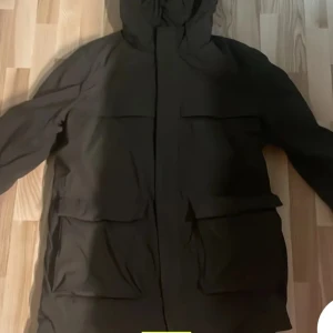 Svart parkajacka med huva - Säljer en stilren svart parkajacka med huva. Jackan har långa ärmar och praktiska fickor framtill. Perfekt för höst och vinter, håller dig varm och skyddad mot väder och vind. Passar både till vardags och lite mer uppklädda tillfällen.