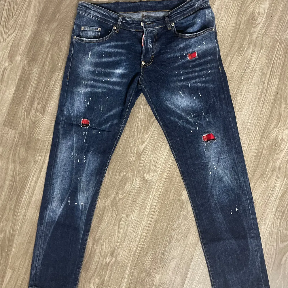 Säljer ett par coola jeans från Dsquared2 i mörkblå denim med slitna detaljer och röda lappar. De har en slim passform och är perfekta för en avslappnad stil. Märkeslappen på baksidan ger en extra touch av stil. Perfekta för höst och vår! Storlek 40. Priset kan diskuteras!. Jeans & Byxor.