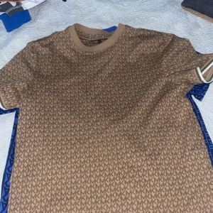 Brun mönstrad t-shirt från Michael Kors - Snygg brun t-shirt från Michael Kors med ett stilrent MK-mönster över hela tyget. Den har korta ärmar med vita och gröna ränder vid ärmsluten. Perfekt för en avslappnad men ändå trendig look. Passar bra till både jeans och shorts!