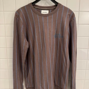 Brun randig tröja från Woodbird i storlek L - Säljer en snygg brun tröja från Woodbird i storlek L. Den är randig med ljusblå ränder och har en broderad text på bröstet. Tröjan är gjord av 100% bomull, vilket gör den superbekväm. Perfekt för både höst och vår!