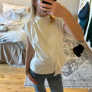 Vit omlottopp - Säljer en stilren vit omlottopp med knytning i sidan. Toppen har en elegant drapering framtill och är ärmlös, vilket gör den perfekt för både vardag och fest. Materialet känns mjukt och bekvämt mot huden. Passar bra till jeans eller kjol för en chic look.