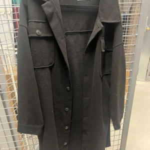 Svart kappa från Nelly  - Säljer en stilren svart kappa från Nelly . Den har en klassisk design med knappar framtill och långa ärmar. Perfekt för höst och vinter, och passar både till vardags och fest. Kappan är i bra skick och redo för nya äventyr!