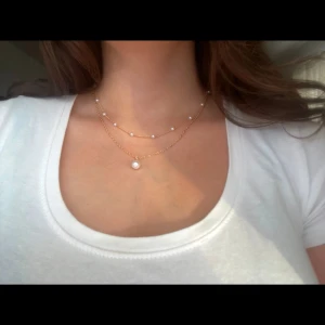 Dubbel halsband med pärlor - Snyggt dubbel halsband  med små vita pärlor. Det övre lagret har flera små pärlor medan det nedre lagret har en större pärla som hänger i mitten. Perfekt för att ge en elegant touch till vilken outfit som helst.