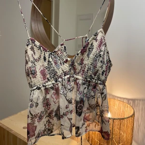 Blommig topp från Zara - Säljer en superfin blommig topp från Zara i storlek XS. Den har smala axelband och ett vackert blommönster i rosa, svart och beige. Toppen är i ett lätt och luftigt material, perfekt för sommardagar. Den har en smockad överdel som ger en skön passform. Perfekt för både vardag och fest! 🌸