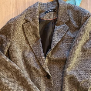 Mönstrad kavaj från Gina Tricot - Snygg kavaj från Gina Tricot i storlek 36. Den har ett klassiskt mönster och är tillverkad i en blandning av polyester och ull. Perfekt för höst och vinter, med långa ärmar och knappar framtill. Passar både till vardag och fest!