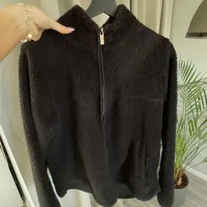 Mysig svart teddyjacka från 157 med dragkedja framtill. Perfekt för kyliga dagar med sin mjuka och fluffiga textur. Jackan har långa ärmar och en hög krage för extra värme. En stilren och bekväm jacka för alla tillfällen.