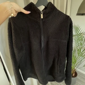 Svart teddyjacka från 157 - Mysig svart teddyjacka från 157 med dragkedja framtill. Perfekt för kyliga dagar med sin mjuka och fluffiga textur. Jackan har långa ärmar och en hög krage för extra värme. En stilren och bekväm jacka för alla tillfällen.