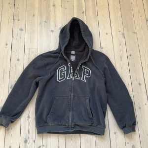 Svart hoodie från GAP - Säljer en svart hoodie från GAP med dragkedja och stor logga på bröstet. Den har en mysig fleecefodrad insida och är tillverkad av bomull och polyester. Perfekt för kyliga dagar med sin bekväma passform och praktiska fickor framtill.