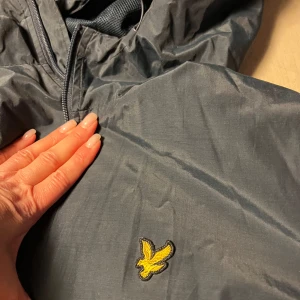 Ljusblå skaljacka från Lyle & Scott - Säljer en stilren ljusblå vind/skal jacka från Lyle & Scott med dragkedja och justerbar huva. Jackan har ett broderat gult emblem på bröstet och är perfekt för blåsiga dagar. Den är lätt och smidig att bära med sig.