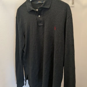 Mörkgrå pikétröja från Ralph Lauren - Säljer en stilren mörkgrå pikétröja från Ralph Lauren med klassisk krage och knappar. Tröjan har långa ärmar och det ikoniska röda logotypbroderiet på bröstet. Perfekt för en avslappnad men ändå elegant look.