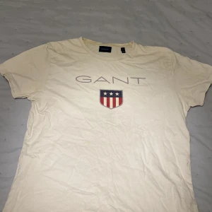 Vit t-shirt från GANT - Säljer en vit t-shirt från GANT med korta ärmar och ett tryck av GANT-loggan och ett sköldemblem på framsidan. Perfekt för en avslappnad stil.