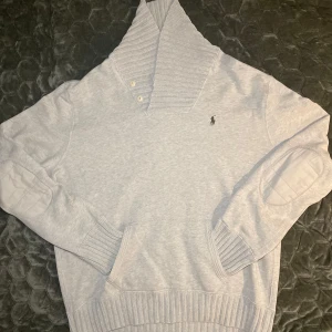 Grå tröja från Ralph Lauren - Säljer en stilren grå tröja från Ralph Lauren med ribbad krage och muddar. Tröjan har en broderad logotyp på bröstet och knappar vid kragen. Perfekt för en avslappnad look. På bild 4 kan man se en fläck på ena under armen, vilket är orsaken till det låga priset