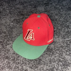 Röd och grön keps med LA-broderi - Snygg röd keps med grönt brätte och broderad LA-logga framtill. Märket Mitchell & Ness är broderat på sidan. Perfekt för att ge en sportig touch till din stil. OBS: ACCEPTERA INTE TILLBAKA SKICKNINGAR