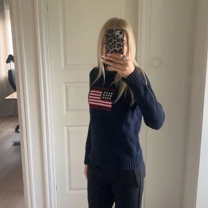 Mörkblå stickad tröja från Polo Jeans Co. - Säljer en mörkblå stickad tröja från Polo Jeans Co. av Ralph Lauren. Tröjan har ett amerikanskt flaggmönster och texten 'Polo Jeans Company' på framsidan. Den är tillverkad i 100% bomull och har långa ärmar med ribbade muddar.