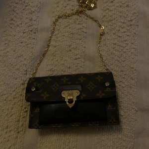Monogram handväska med kedja - Snygg handväska från Louis Vuitton med klassiskt monogrammönster. Väskan har en guldfärgad kedja och ett elegant lås framtill. Perfekt för att ge din outfit en lyxig touch.