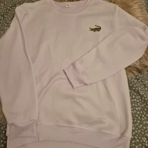 Vit sweatshirt från Lacoste - Säljer en stilren vit sweatshirt från Lacoste med det klassiska krokodilemblem på bröstet. Tröjan har långa ärmar och ribbade muddar vid ärmslut och nederkant. Perfekt för en avslappnad look.