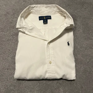 Vit skjorta från Ralph Lauren - Klassisk vit skjorta från Ralph Lauren med broderad logotyp på bröstet. Skjortan har långa ärmar och knappar framtill. Perfekt för en stilren look. Stl xs