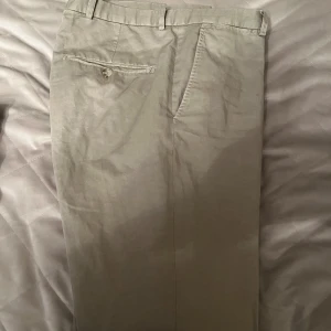 Beige chinos från J.Lindeberg - Snygga beige chinos från J.Lindeberg i slim fit. Tillverkade i en skön blandning av bomull och linne, perfekta för en stilren look. Byxorna har knappar och bälteshällor för extra stilpoäng.