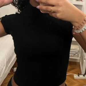 Svart ribbad polotopp - Säljer en stilren svart ribbad polotopp med korta ärmar. Perfekt för en chic och enkel look. Toppen har en tight passform och är gjord i ett mjukt material som känns skönt mot huden.