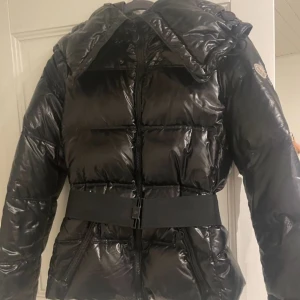 Svart pufferjacka från Moncler - Snygg svart pufferjacka från Moncler med glansig finish och bälte i midjan. Jackan har en dragkedja framtill och långa ärmar. Perfekt för kyliga dagar när du vill hålla stilen.