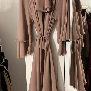 Abaya - Elegant abaya med knytband. Abayan har en mjuk och flödande design med subtila volangdetaljer vid ärmsluten. Perfekt för en fest. Matchande sjal följer med. Användes endast en gång. (Kan fraktas)