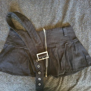 Svart kortkjol med bälte - Snygg svart riktigt mini kortkjol med dragkedja framtill och ett brett bälte med metallspänne. Kjolen har en stilren design och passar perfekt för en trendig look. Pälsen rollas bort vid försäljning 