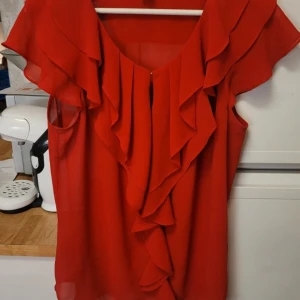 Röd blus med volangdetaljer - Säljer en elegant röd blus med vackra volangdetaljer framtill. Blusen har en luftig design och är perfekt för att ge en färgklick till din garderob. Passar bra till både jeans och kjol.