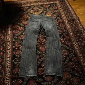 Blå jeans från Guess - Blå jeans från Guess med klassisk design och en diskret logga på bakfickan. Jeansen har en rak passform och är perfekta för en avslappnad stil. Sömmarna är i mörkblå färg och jeansen har en knappgylf.