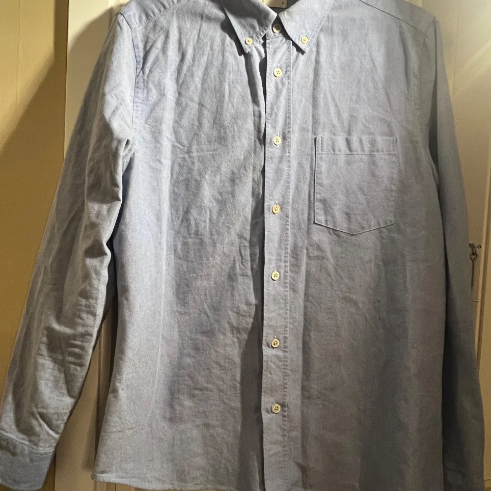 Säljer en stilren blå Oxford-skjorta från Asket i storlek S. Skjortan har en klassisk krage med knappar och en bröstficka. Perfekt för en avslappnad men ändå uppklädd look. Tillverkad i bomull för en bekväm passform.. Paidat.