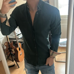 Burberry skjorta - Snygg grön skjorta med långa ärmar och stiliga rutiga detaljer på insidan av ärmsluten. Perfekt för en avslappnad men ändå uppklädd look. Skjortan har knappar framtill och en bröstficka. Liten defekt på fjärde bilden