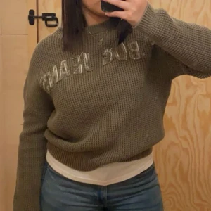 Olivgrön stickad tröja från BDG - Säljer en olivgrön stickad tröja från BDG med texten 'BDG JEANS' på framsidan. Tröjan har en avslappnad passform och långa ärmar, perfekt för kyligare dagar. Den är gjord i ett grovt stickat material som ger en mysig känsla. Pris kan diskuteras 