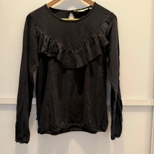 Svart långärmad topp från House of Lola - Snygg svart långärmad topp från House of Lola i mjukt tencel-material. Toppen har en volangdetalj framtill och en liten öppning i nacken. Perfekt för en stilren look.