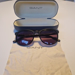 Svarta solglasögon från Gant - Snygga svarta solglasögon från Gant med runda glas och mörka linser. Kommer med ett praktiskt fodral och putsduk. Perfekta för en stilren look.