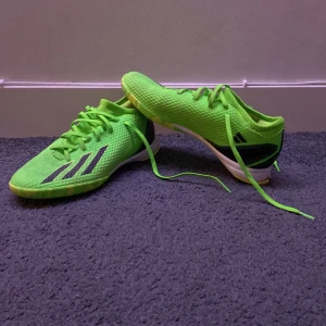 Gröna fotbollsskor från Adidas - Snygga gröna fotbollsskor från Adidas med svarta detaljer och snörning. Skorna har en mönstrad sula för bra grepp och är perfekta för inomhusspel. De har en modern design med tre svarta ränder på sidan.