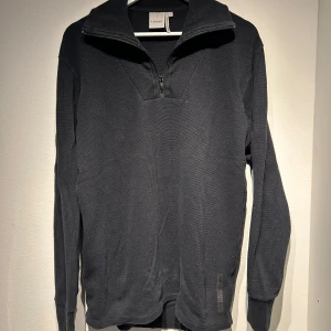 Svart tröja med dragkedja från Elvine - Snygg svart tröja från Elvine med dragkedja vid halsen. Tröjan har en ribbad struktur och långa ärmar, perfekt för kyligare dagar. Enkel och stilren design som passar till det mesta.